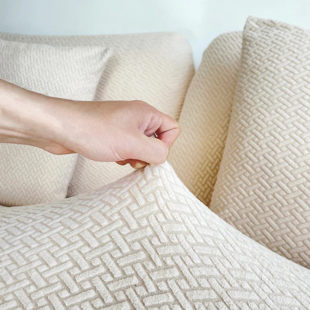 Close-up of a hand gently pinching and lifting the edge of a cream-colored textured pet-friendly sofa cover on a cushion, highlighting the soft, woven herringbone pattern fabric against a light background with additional covered cushions in view.