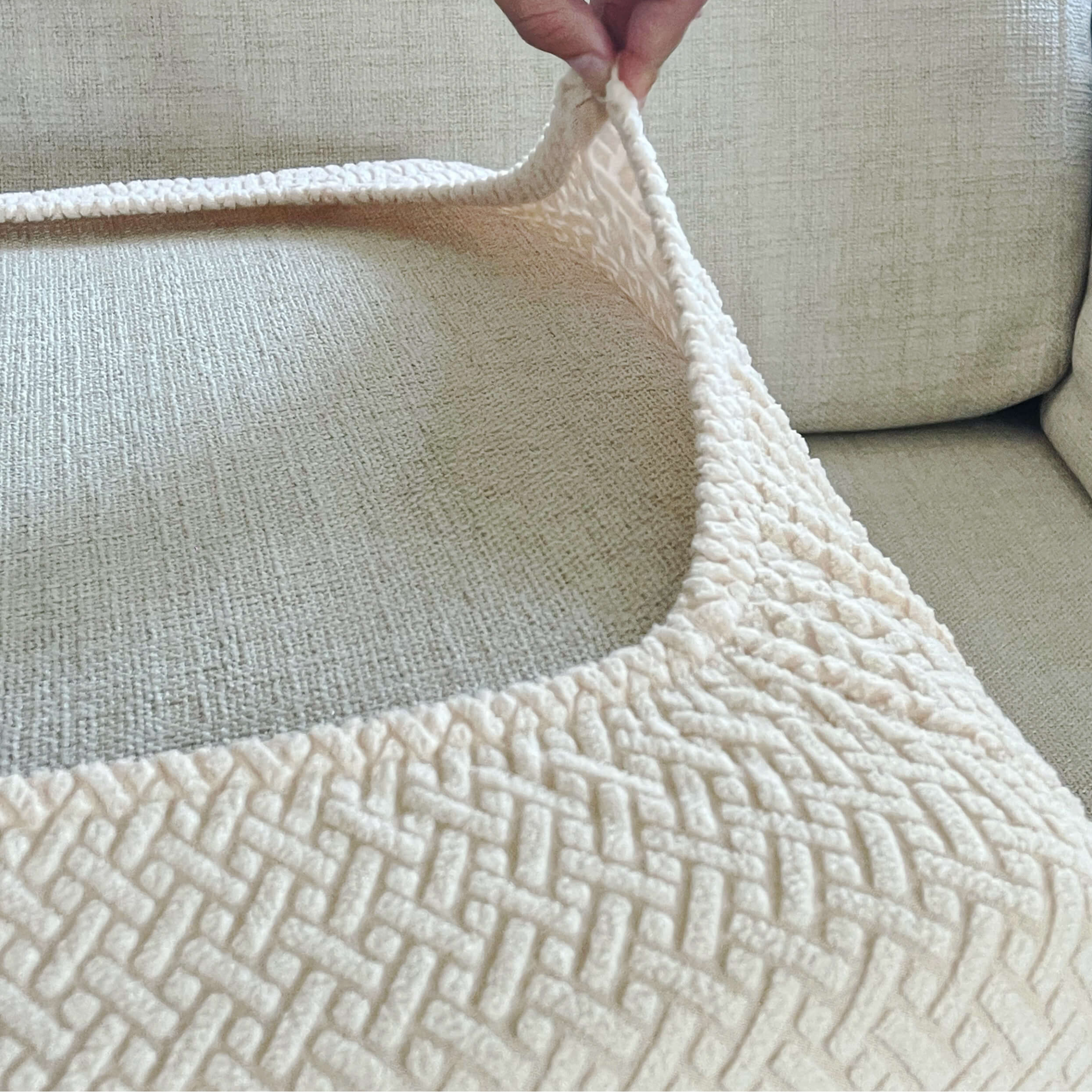 Close-up of a hand lifting the corner of a white textured pet-friendly sofa cover fitted on a beige couch cushion, demonstrating the elastic fit and soft, woven herringbone pattern fabric.